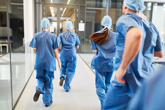 Rennendes Ärzteteam in blauer OP-Kleidung auf Krankenhausflur von hinten